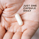 Hand holding a white capsule with text 'JUST ONE CAPSULE DAILY' on a blurred background