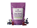 A purple stand-up pouch of True Sea Moss Sea Moss Elderberry Gummies, styled with fresh elderberries and red gummy bears on a light lavender background.